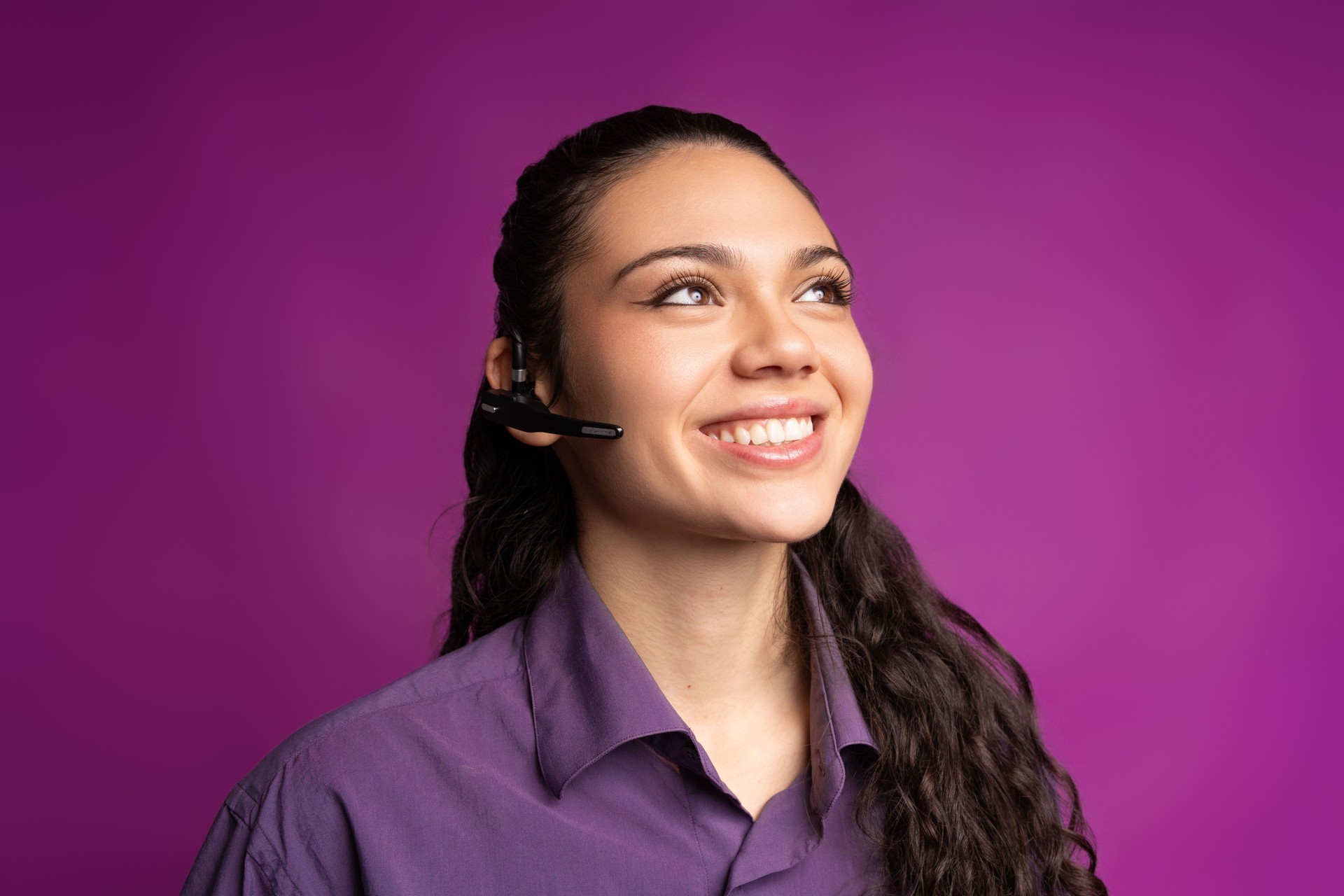 Portrait of a cheerful young woman wearing a headset, looking up and smiling on a vibrant purple background